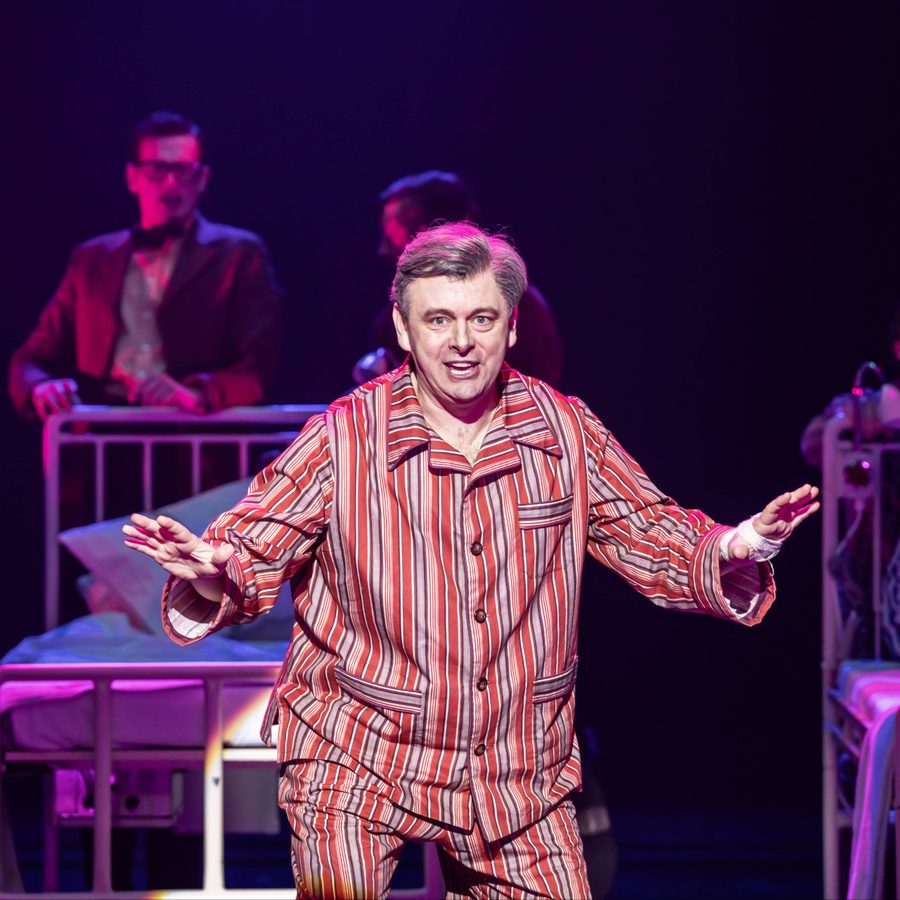 Michael Sheen wearing red striped pyjamas. He is mid-song. The ensemble cast move hospital beds behind him.