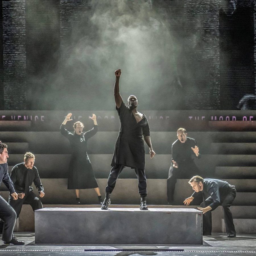 Othello stands centre stage on a raised platform, his fist raised, surrounded by figures in expressive poses.