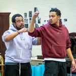 Two actors in a rehearsal room. One is wearing a light purple hoodie and glasses, using a tape measure to measure the length of the other actor's arm.