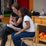 Two young people sitting on chairs looking at a script. They are holding a phone between them. In the background another young person watches them, smiling.