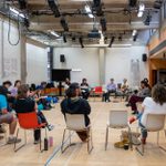 A group of young people sitting in a circle on chairs ina rehearsal room.