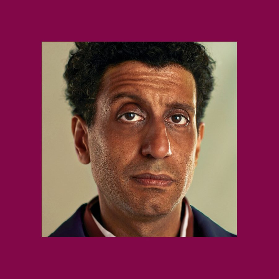 A man with dark curly hair gazes upward with a thoughtful expression. He wears a dark suit jacket over a shirt. The background is a muted, light color, and the image is centered on a maroon backdrop.