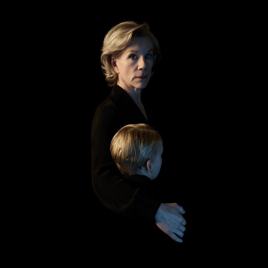 A woman with short blonde hair, wearing a dark outfit, looks intently at the camera. In front of her is a child with blonde hair whose face is turned away from the camera. They are set against a black background.