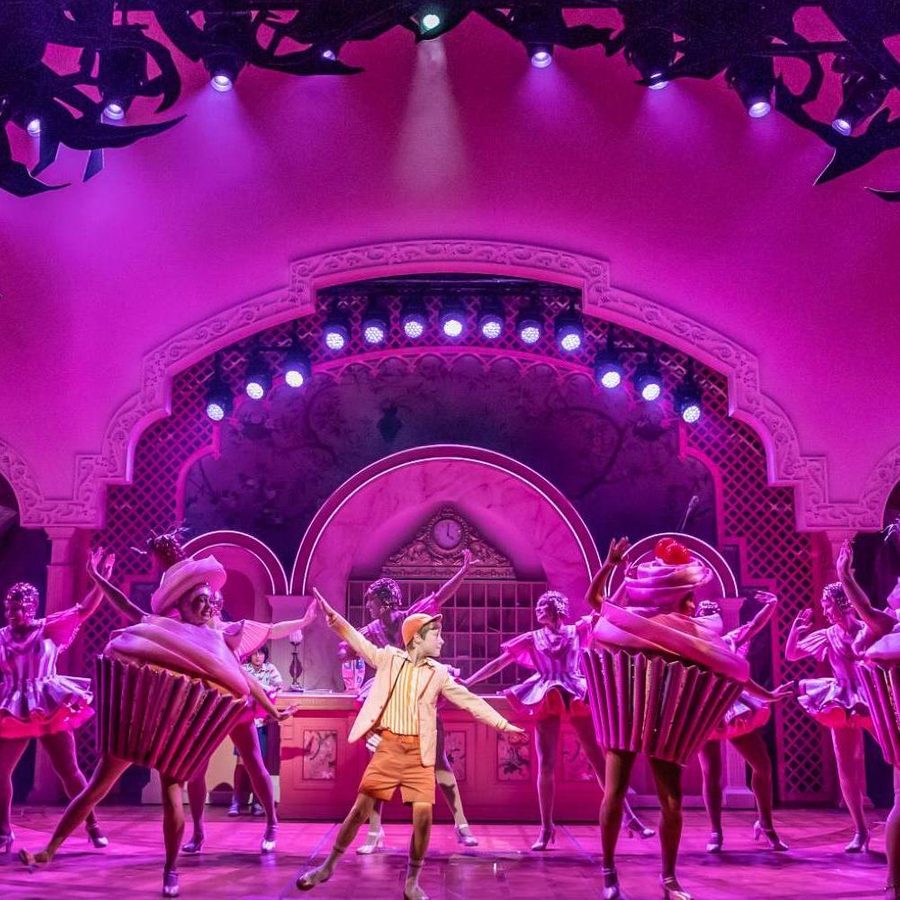 A spotlight picks out a young boy wearing shorts, jacket and cap, dancing in the centre of a line up of dancing cup cakes, with another row of dancers behind.