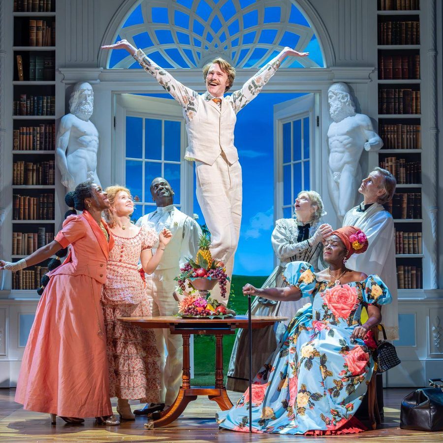 A group of seven actors on stage in the set of a grand house. The actor in the centre is stood on a circular wooden table with his arms up in a joyful pose. The other actors are around him looking up at him with varying smiles or frowns.