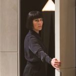 A person with a short, dark bob haircut stands partially behind a door. They are wearing a long-sleeved, dark outfit and looking directly forward, with one hand on the edge of the door.
