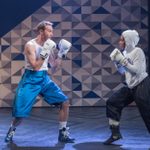 Two people wearing boxing gloves face off centre stage. The boxer on the left wears blue shorts and a white tank top. The boxer on the right wears a white hoodie and black trousers.