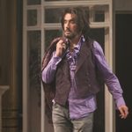 A person with long hair and a beard, wearing a purple shirt and waistcoat, and holding a coat over their shoulder. They are scruffy looking.