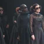 A group of people stand on stage, dressed in dark, formal outfits and sunglasses. They are all looking in the same direction.