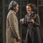 Two people face each other. One wears a herringbone coat and cap, while the other is in a dark vintage dress, holding a banknote. Both are smiling, with a backdrop of windows and geometric patterns.