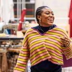 A woman in a yellow and pink striped jumper, laughing and looking to her left. Behind her is a table with a sewing machine and various clothes hung on railings.