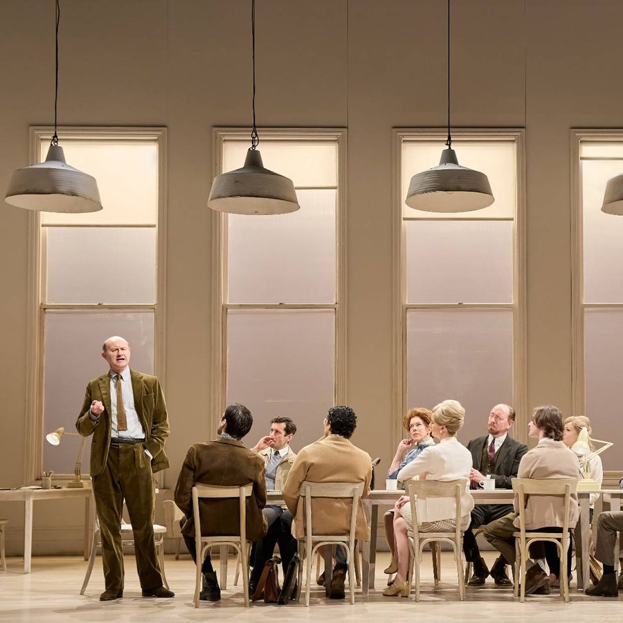 Mark Gatiss is gesturing with his hand. He wears a brown-green corduroy suit, white shirt, black belt and brown shoes. A group of people sit around a long table to the right of the man, listening to him intently.