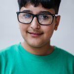 A young person with short black hair and glasses is wearing a green T-shirt, smiling subtly at the camera against a plain background.