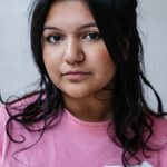 A young person with long dark hair and a pink shirt looks directly at the camera. The shirt has the words 