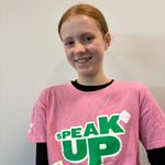 A person with red hair smiles at the camera. They are wearing a pink t-shirt with the words 