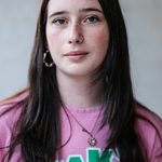 A person with long dark hair stands against a plain background, wearing a pink shirt with green and white text. They have hoop earrings, a nose ring, and a necklace, and they are looking directly at the camera.