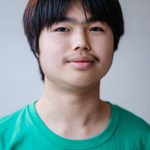 A young person with short black hair and a faint mustache, wearing a green T-shirt, stands against a plain background. They have a neutral expression and face the camera directly.