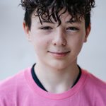 A young person with curly dark hair is wearing a pink T-shirt and a black underlayer, standing in front of a plain light background, smiling slightly at the camera.