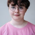 A person with short, curly brown hair and glasses smiles gently at the camera. They are wearing a pink t-shirt with green and white lettering. The background is plain and light-colored.