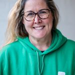 A person with long hair and glasses smiles while wearing a bright green hoodie with the words 