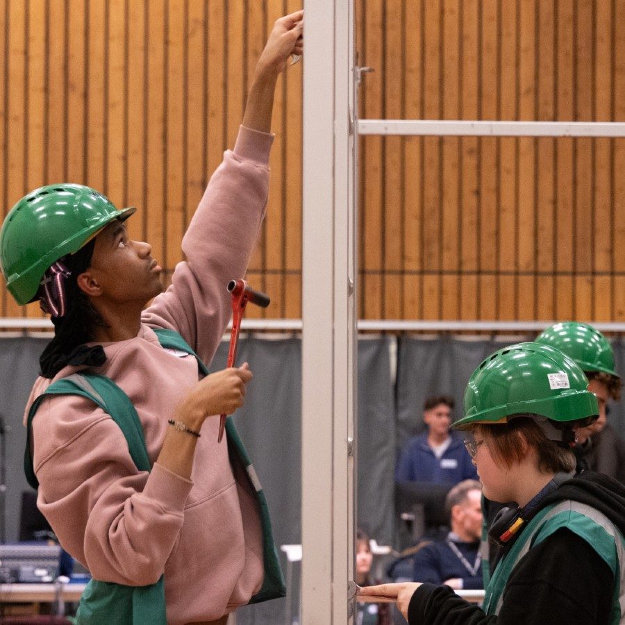 A group of young people wearing green safety helmets and green hi-vis jackets work on assembling a metal frame. They are indoors, with wood panel walls in the background.