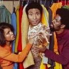 Three people are playfully interacting among colorful clothes hanging on a rack. The person in the center is being playfully grabbed by the others, creating a humorous scene against a backdrop of various vibrant garments.