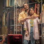 An actor and an actress stand closely in front of a railing filled with colourful 1970s clothes. The actor wears a yellow shirt, and the actress wears an oversized pink shirt.