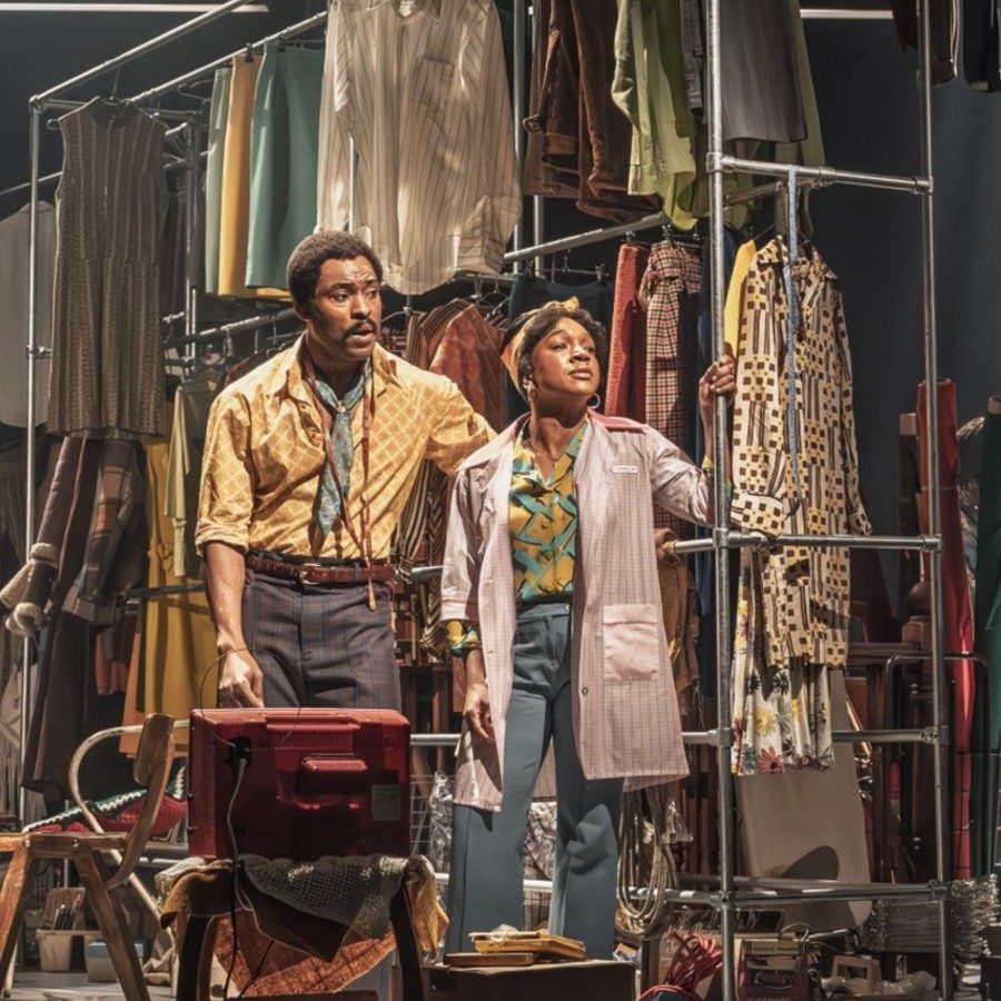 An actor and an actress stand closely in front of a railing filled with colourful 1970s clothes. The actor wears a yellow shirt, and the actress wears an oversized pink shirt.