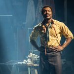 An actor stands confidently on a stage, illuminated by a spotlight. They wear a yellow patterned shirt, brown plaid pants, and a blue scarf. Behind them is a table with sewing equipment, and clothing hangs in the background.