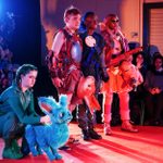 Four performers in colorful, dystopian-themed costumes stand on a stage, with one crouching beside a blue, rabbit-like puppet. An audience of seated spectators watches from the sides.