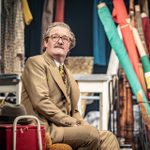 An actor in a tan suit and glasses sits pensively on stage surrounded by colourful fabric rolls and props.