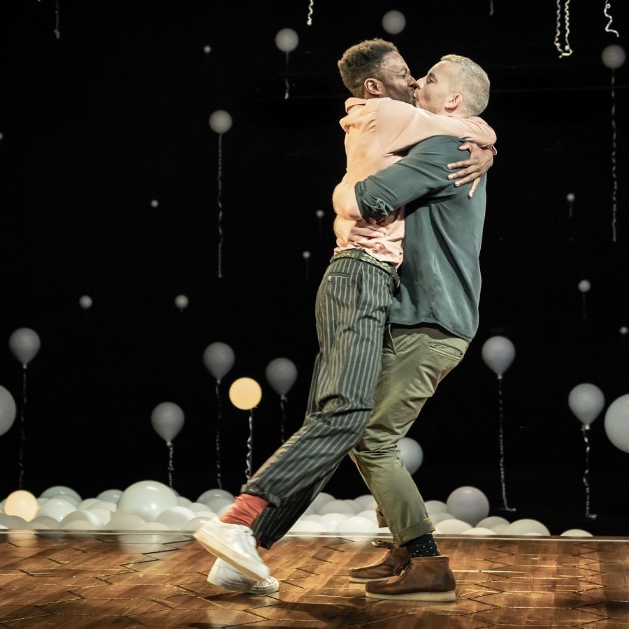 Two men are joyfully embracing and kissing on a stage decorated with white balloons. One person lifts the other as they share a moment of celebration. They are surrounded by a dark backdrop with hanging strings and scattered balloons on the floor.