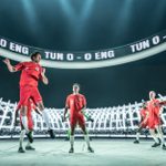 Seven soccer players in red uniforms are on a brightly lit stage with a digital backdrop displaying 