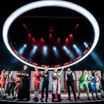 A diverse ensemble of performers stands on stage under a circular light, dressed in vibrant costumes. A digital display above shows the score 