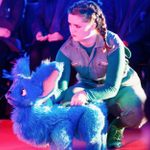 A person with braided hair crouches beside a large, blue furry puppet with big ears. They are both looking the same way.