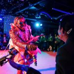 A performer in a colourful, eccentric costume smiles and extends hand towards a seated audience member. They're holding a large gun and are surrounded by bubbles on a stage with vibrant pink lighting.