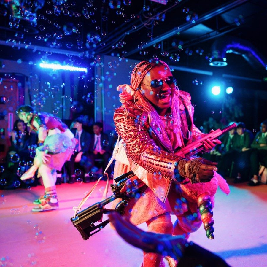 A performer in a colourful, eccentric costume smiles and extends hand towards a seated audience member. They're holding a large gun and are surrounded by bubbles on a stage with vibrant pink lighting.