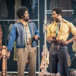 Two actors stand in a cluttered tailor shop with various clothing items and fabric in the background. The actor on the left wears a blue cardigan and brown trousers. The actor on the right wears a yellow patterned shirt and a blue scarf.