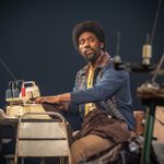 An actor sits at a sewing machine, surrounded by sewing supplies. They are wearing a blue cardigan and brown trousers.