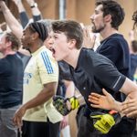 A group of people enthusiastically cheers and gesturing, suggesting excitement or encouragement. One person wearing goalie gloves is shouting energetically, while others clap and raise their arms.