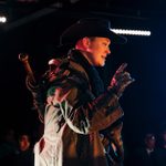 A performer dressed in a cowboy hat, coat and leather gloves is standing on stage, looking down with one raised finger.