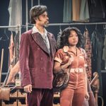 Two actors on stage with various vintage clothing items hang on racks. The man wears a maroon velvet suit, and the woman is in an orange jumpsuit holding a handbag.