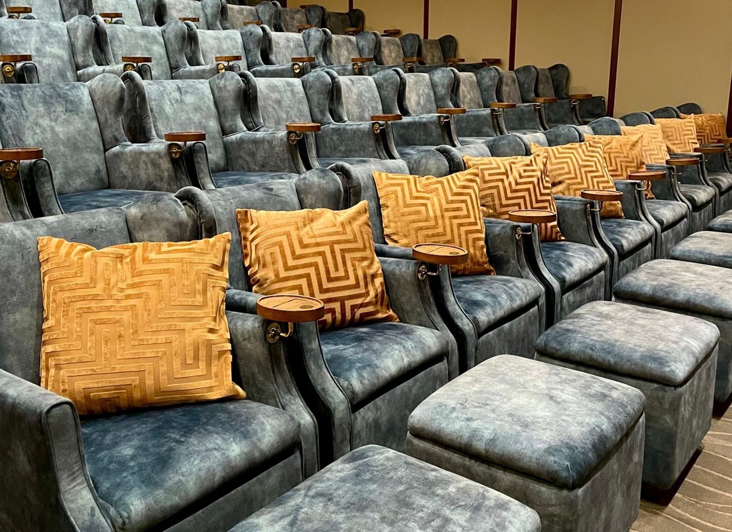 A theatre with rows of plush, blue/grey seats featuring wooden armrests and cup holders. Each seat has a large, patterned gold cushion. Matching blue footrests are in front of each seat. The floor is carpeted in a neutral tone.