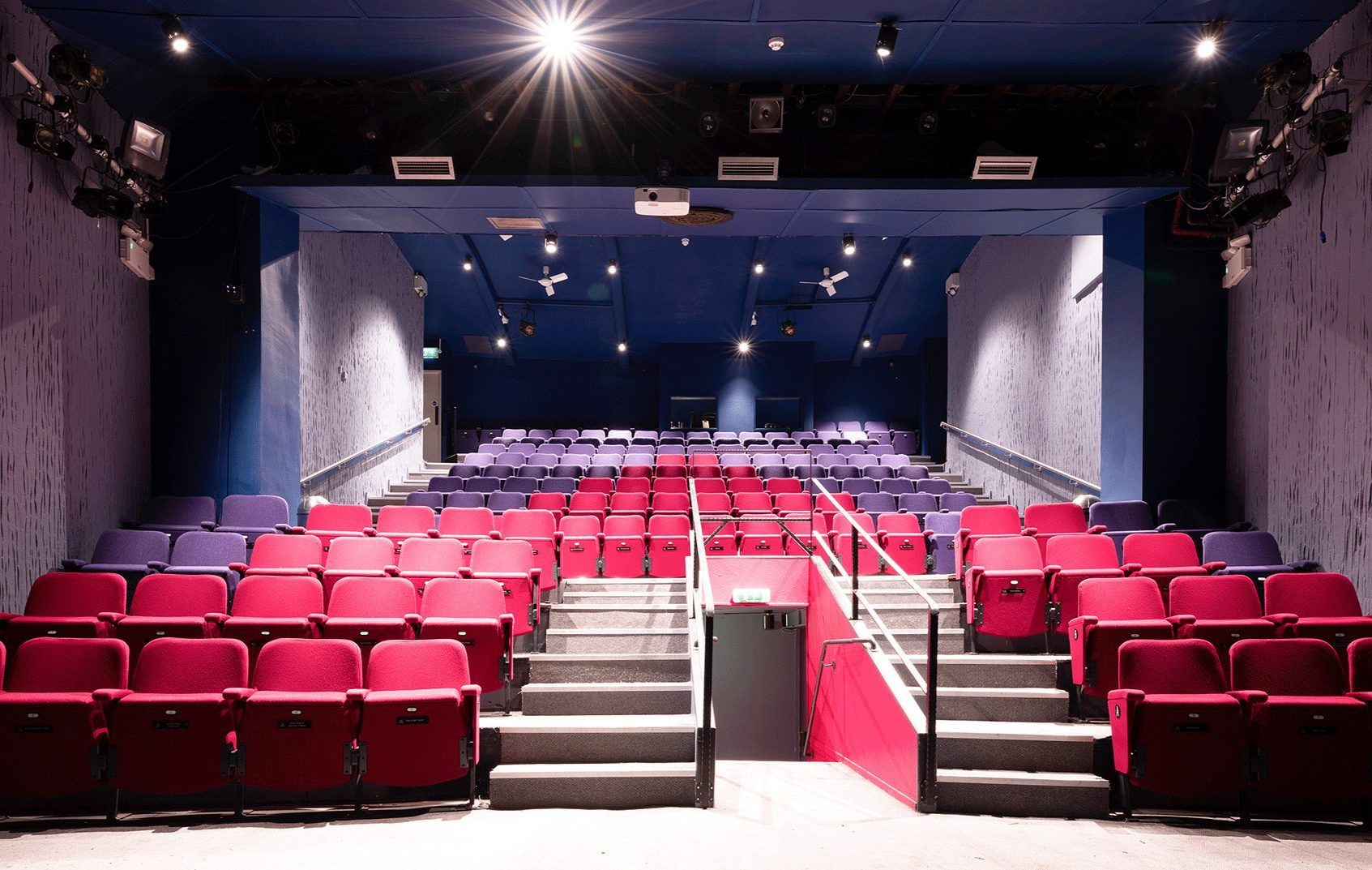 Interior view of a modern theatre with red and purple seats arranged in tiered rows. Stairs with handrails divide the seating sections. Ceiling lights illuminate the space, highlighting the carpeted floor and neutral-toned walls.