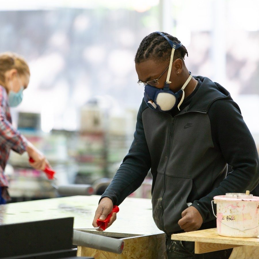 Two young people are in a workshop. They are painting pieces of scenery using paint rollers. A tub of paint is next to them with more scenery in the background of the workshop.