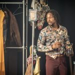 An actor holding a coffee pot, wearing a patterned shirt and brown pants. On his right are clothing racks with various colourful clothes hanging.