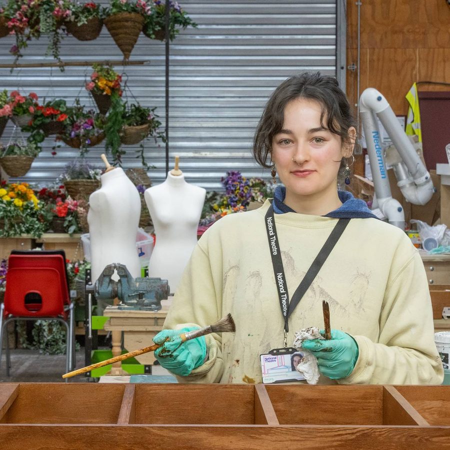 A person stands in a workshop holding a paintbrush, wearing gloves and a lanyard. Behind them are mannequins, vibrant flower baskets, and various tools. A wooden shelving unit is in front of them. The setting is busy and creative.