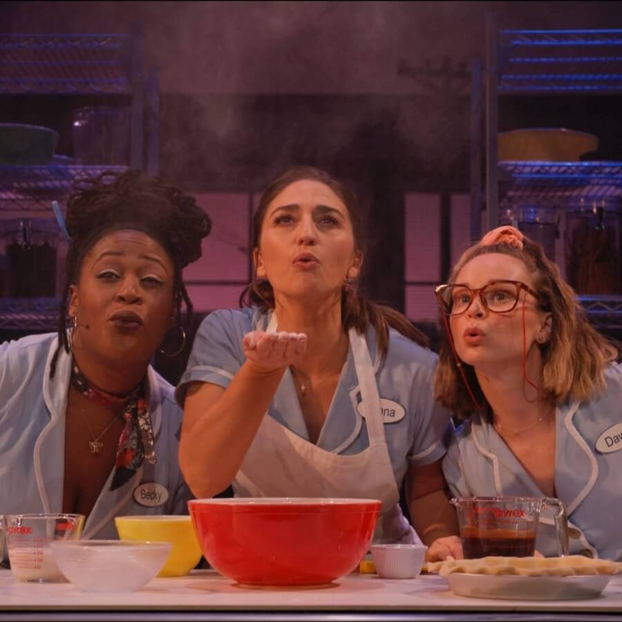 Three women in diner uniforms blow flour toward the camera from a kitchen counter. They are surrounded by baking ingredients, including a red mixing bowl and a measuring cup. The background reveals shelves with various kitchen supplies.