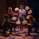 A woman in a nurse outfit stands surrounded by four people kneeling and holding different pies around her. The scene is dimly lit, creating a theatrical atmosphere on a checkered floor.