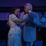 A woman in a light dress and an older man in a light suit with a bow tie dance together on stage. The scene is dimly lit with a purplish hue. Other performers are visible in the background.